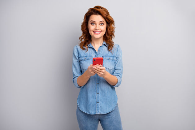 社交兴奋的正面开朗的女人的肖像使用手机喜欢发短信打字博客帖子穿着好看的衣服隔离在灰色的墙上红发卷发博客