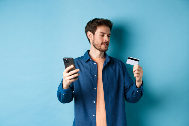 男性电子商务概念男人在线支付 读信用 拿着手机 站在蓝色的背景上胡须发型移动