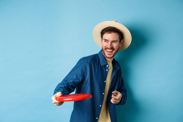 胡子快乐的男人笑着玩飞盘 朝朋友扔东西笑着 站在蓝色背景的夏日帽子里年轻脸男