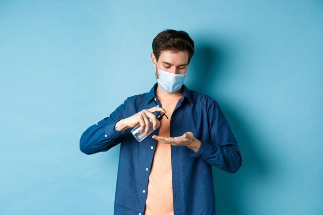 男人Covid-19 健康和流行病概念年轻人戴着医用口罩用消毒液洗手 在手掌上涂上防腐剂 站在蓝色的背景上英俊朋友年轻