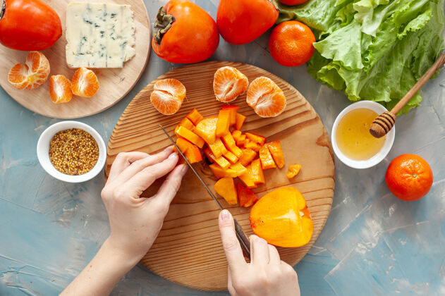 吃用柿子 橘子和蓝奶酪做维生素冬季沙拉的原料和过程一步一步地那个女人在一块圆木板上切柿子从上面看素食俯视图烹饪