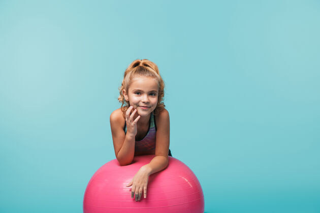 微笑快乐的小女孩穿着运动服 靠在一个健身球上 隔着蓝色的墙小活跃健身