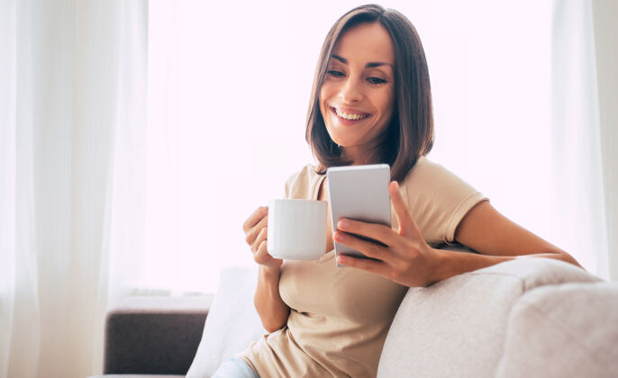 无线年轻的现代自信微笑的女人在家工作或放松 坐在沙发上用智能手机使用信息漂亮