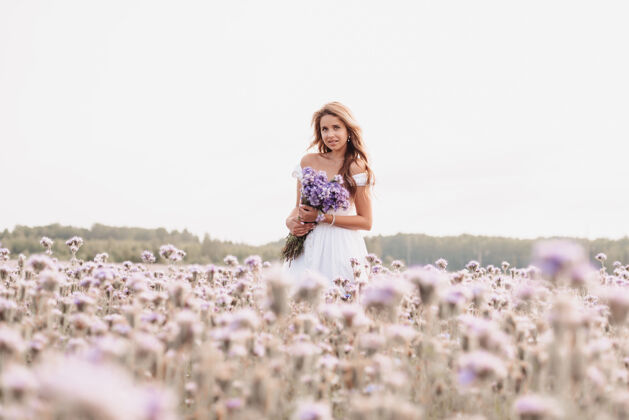 衣服美丽的年轻女子 穿着白色的连衣裙 手拿一束鲜花 在大自然夏日的田野里白天放松花