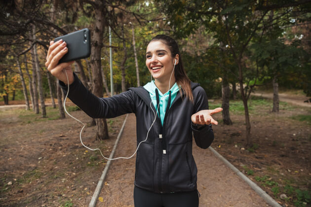 运动微笑的健身女士戴着耳机听音乐 站在公园里自拍休息深色头发运动