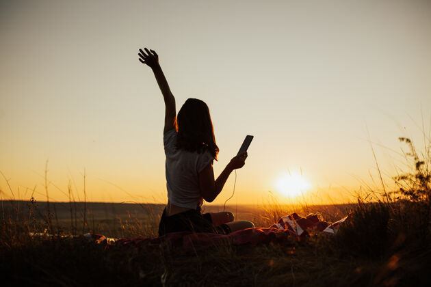 阳光美丽的夕阳下 女孩坐在球场上 享受着在夕阳下用带耳机的手机应用听音乐日落微笑山