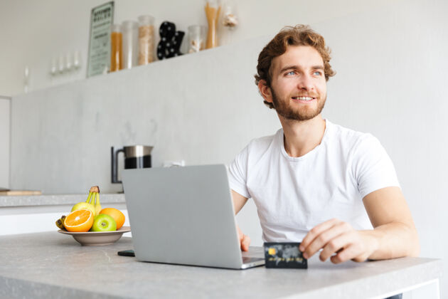 年轻快乐的小胡子男人在家里的桌子旁用手提电脑拿着信用卡聊天放松舒适成人