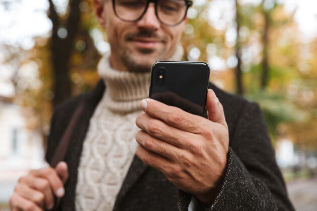 站立30多岁的帅哥穿着暖和的衣服 在户外穿过秋天公园 并使用智能手机的照片天气公园帅气