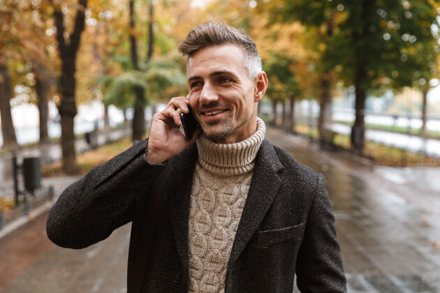 电话一张30多岁的白种人穿着暖和的衣服 在户外穿过秋天的公园 并使用手机的照片休闲站立寒冷
