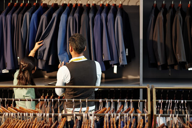 选择首席裁缝和工作室经理检查挂在栏杆上的定制夹克的质量 从后面看货架质量销售助理