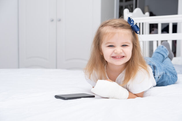创伤手拿石膏的小女孩躺在床上用智能手机 看动画片或教育视频童年移动事故