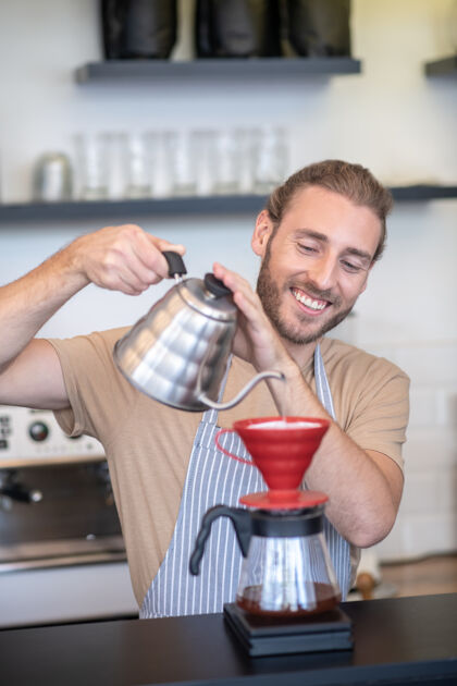 倒酿造咖啡微笑围裙上的男人站在咖啡馆里往茶壶里倒水冲咖啡过滤器好看过程