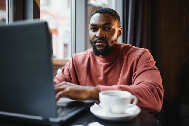 电脑快乐的非洲商人坐在咖啡馆和笔记本电脑工作的肖像互联网咖啡工人