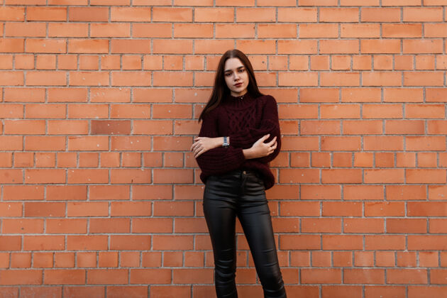 皮革身穿紫红色毛衣和黑色皮裤的年轻女子站在城市的砖墙附近女孩毛衣美女