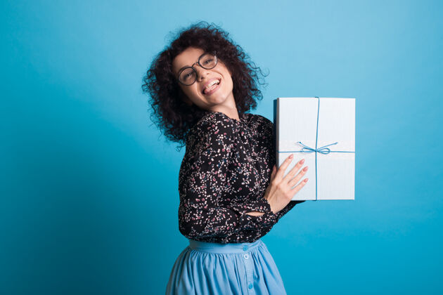 持有一位戴着眼镜的卷发女士穿着一件连衣裙 在蓝色的工作室墙上摆着一个盒子 里面摆着礼物盒子女人单色