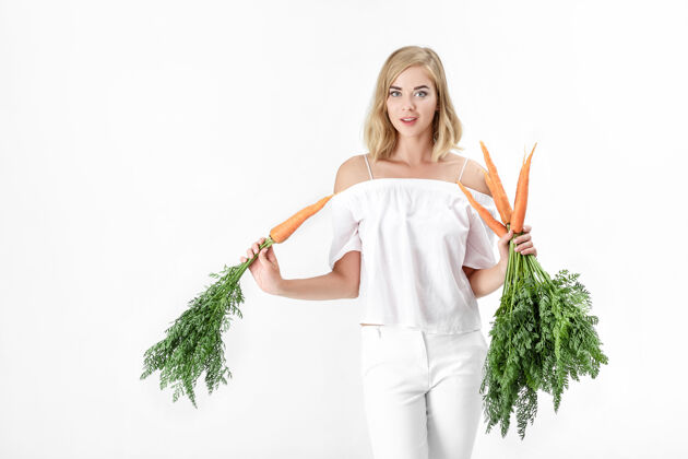 营养一个穿着白衬衫的金发美女 手里拿着一根新鲜的胡萝卜 上面有绿叶背景.健康还有节食束手维生素