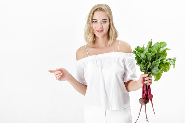 蔬菜一位穿着白色上衣的金发美女手持一根长着绿叶的甜菜根背景.健康还有维生素植物女人微笑