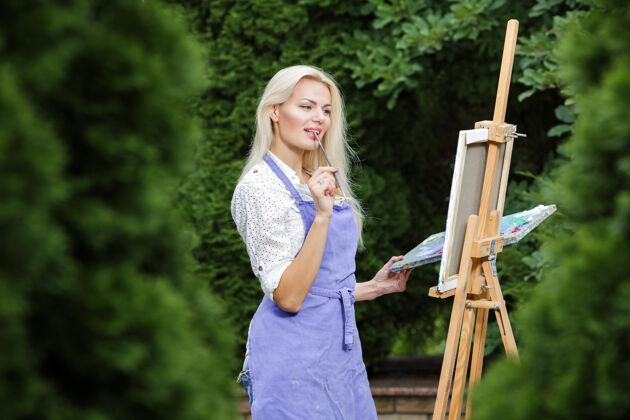 幻想美丽的金发女画家手里拿着画笔在花园里的画布上画画金发女郎女性人物