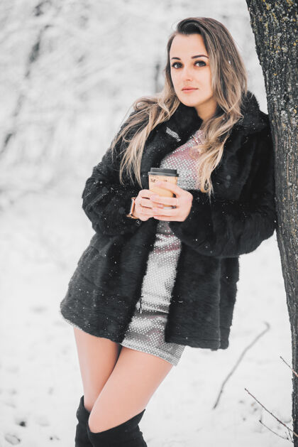 女孩冬天在森林里 一个穿着皮衣长裙和高筒靴的漂亮女孩手里拿着咖啡性感心情时尚