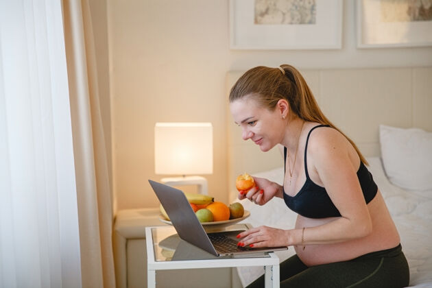 金发一个年轻的金发孕妇坐在床上 一边吃水果一边看着她笔记本电脑高高质量的照片看盘子演习