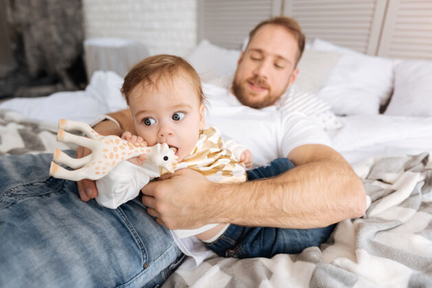 可爱最喜欢的玩具高兴包括细心的父亲躺在家里的床上 抱着蹒跚学步的女孩 而他的孩子在玩玩具父亲身份发展新生儿
