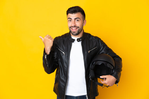 皮革高加索男子戴着摩托车头盔 在孤立的黄色背景下指着侧面展示产品拉丁语年轻人自信