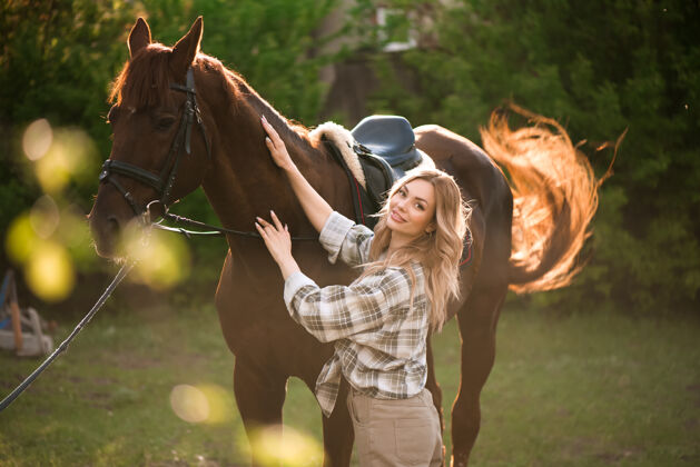 骑手美丽的姑娘牵着她的马和美丽温暖的夕阳在春天的森林里公园马头发