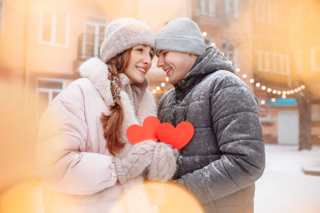 情人冬日里 一对年轻的情侣捧着红色的纸心 在外面感受着快乐和幸福爱情浪漫男人和女人在公园的雪下庆祝情人节羊毛快乐情侣