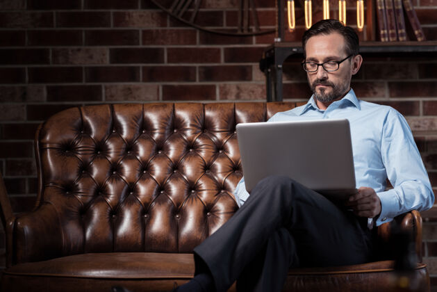 现代使用技术认真的勤劳的个体户坐在沙发上 膝盖上放着一台笔记本电脑 一边工作工作数码男士