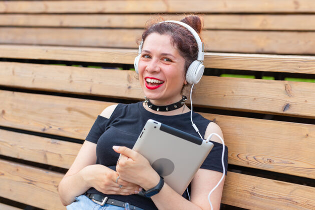 女性一位美丽时尚的年轻开朗女子的肖像戴着耳机坐在公园里取暖肖像哥特朋克