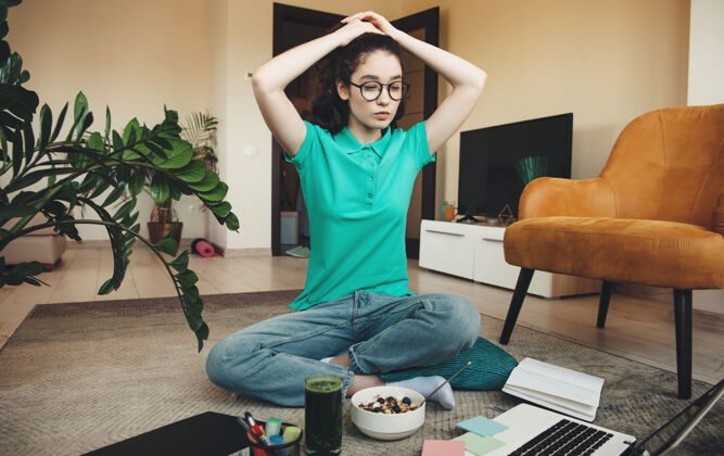 大学一个戴着眼镜的忙碌的白人妇女在地板上做着疲倦的手势 一边用笔记本电脑上课 一边用新鲜果汁吃麦片笔记笔记本电脑阅读