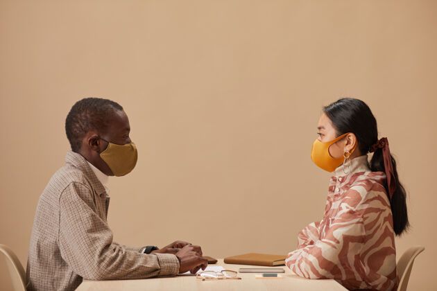 防疫两个戴着防护面具的同事坐在桌子旁 在会议期间互相看着对方的侧视图防护面罩职业商务女性
