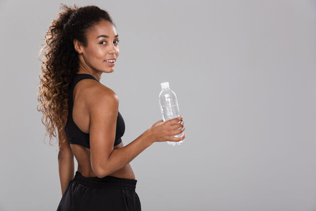 体重一位快乐的年轻非洲女运动员的肖像 她在灰色的墙上独自训练后休息 手里拿着装满水的瓶子健身房饮食活力