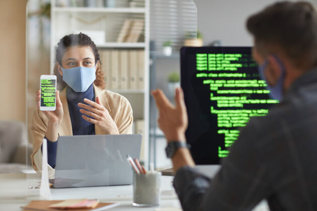 编码戴着面具的女人坐在同事对面的桌子旁 一边在办公室工作一边给他看带着软件的手机团队技术长相