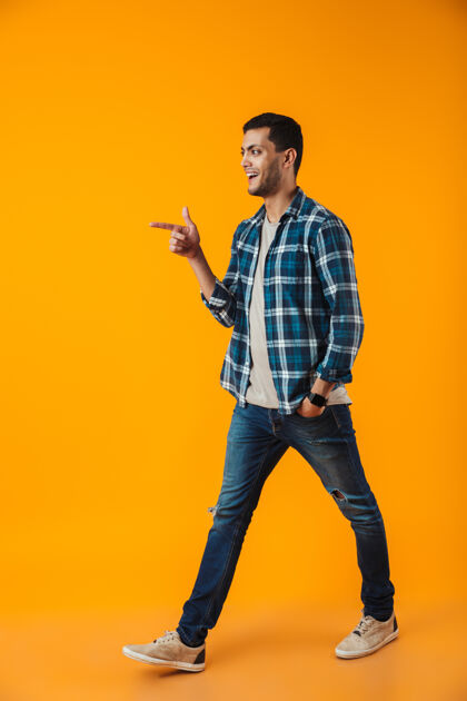 男人一个穿着格子衬衫的快乐的年轻人的全长肖像被隔离在橙色的墙上 走着 指着别处好时尚成人