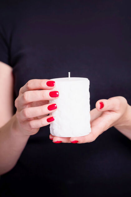 黑色衣服美丽的女性双手握着白色蜡烛修指甲用红色指甲油抛光女性蜡烛