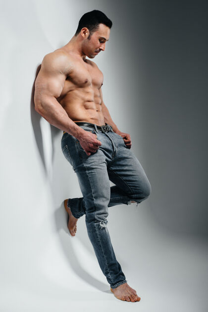 健美一位年轻的健美运动员赤裸上身 穿着牛仔裤站在墙边的摄影棚里运动身体适当的营养力量