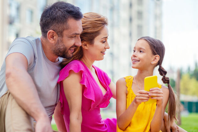 女孩好心情积极快乐的女孩看着父母 手里拿着智能手机家庭日照顾关系