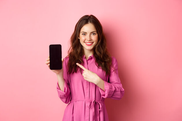 休闲年轻的白种女人穿着时髦的衣服 手指着智能手机屏幕 微笑着 在网上展示促销交易 粉色的墙壁请黑发惊人