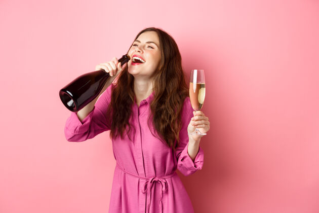 惊人庆祝和节日的概念快乐无忧无虑的女人喝着香槟酒瓶高兴的微笑 放松在聚会上 拿着杯子 站在粉红色的墙上女性情绪积极