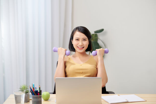 亚洲人微笑的孕妇坐在椅子上 手里拿着两个哑铃 看着笔记本电脑笔记本电脑自由职业者女性