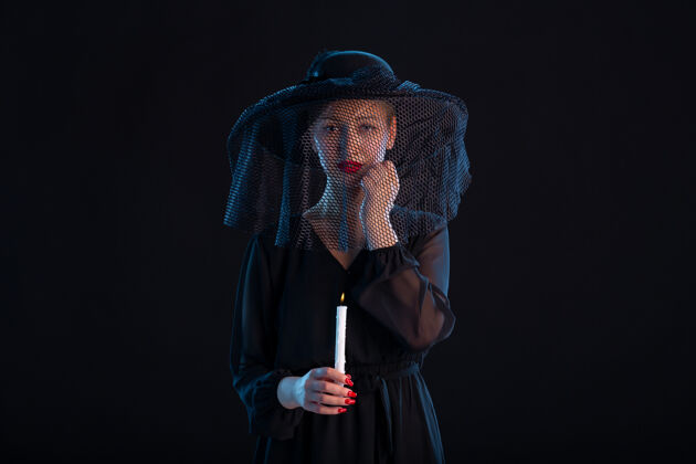 表情悲哀的黑衣女子用燃燒的蠟燭對黑色悲傷葬禮死亡帽子火災蠟燭