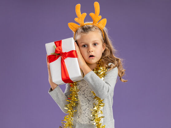 小惊讶的小女孩戴着圣诞发箍 脖子上戴着花环 脸上挂着礼品盒 孤零零地贴在蓝色的墙上脖子花环举行