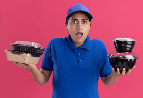 姿势困惑的年轻送货女孩穿着制服 戴着帽子 把食物容器隔离在粉红色的墙上衣服食物人