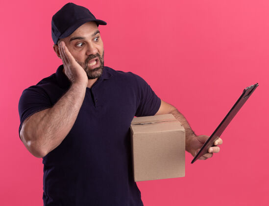 衣服惊恐的中年送货员穿着制服 拿着帽子盒 看着他手里的剪贴板孤立在粉红色的墙上帽子手姿势