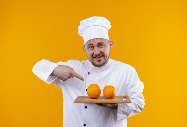 印象穿着厨师制服的年轻帅哥拿着砧板 上面放着橘子 指着隔离在橘色墙上的橘子年轻厨师橙色