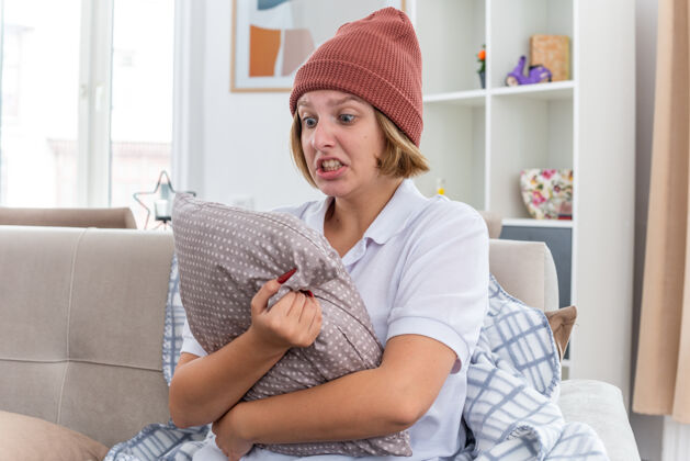 痛苦愤怒的不健康的年轻女子 戴着暖和的帽子 穿着毛毯 看起来不舒服 生病 患感冒和流感 抱着枕头 表情咄咄逼人 坐在客厅的沙发上温暖侵略性愤怒