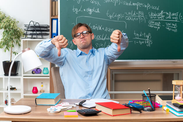 穿戴眼镜的年轻男老师不高兴地竖起大拇指坐在课桌旁 教室的黑板前放着书和笔记笔记不高兴拇指