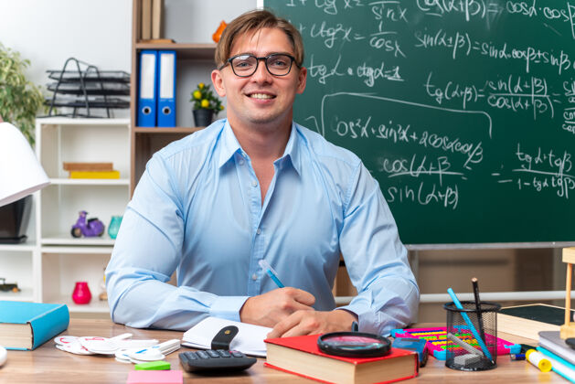 年轻快乐微笑的年轻男老师戴着眼镜坐在课桌旁 教室的黑板前放着书和笔记课桌教室笔记