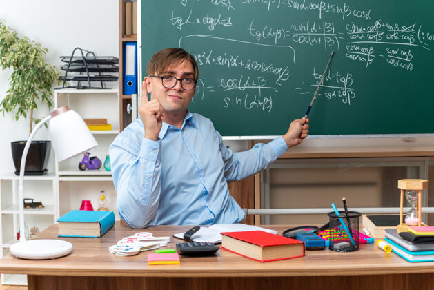 指针年轻的男老师戴着眼镜 带着教鞭 自信地坐在课桌旁 在教室的黑板前放着书和笔记黑板少年笔记